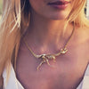 Woman wearing a yellow-gold Tyrannosaur NEX necklace—an articulated T-rex skeleton draped across her collarbone in warm summer light.