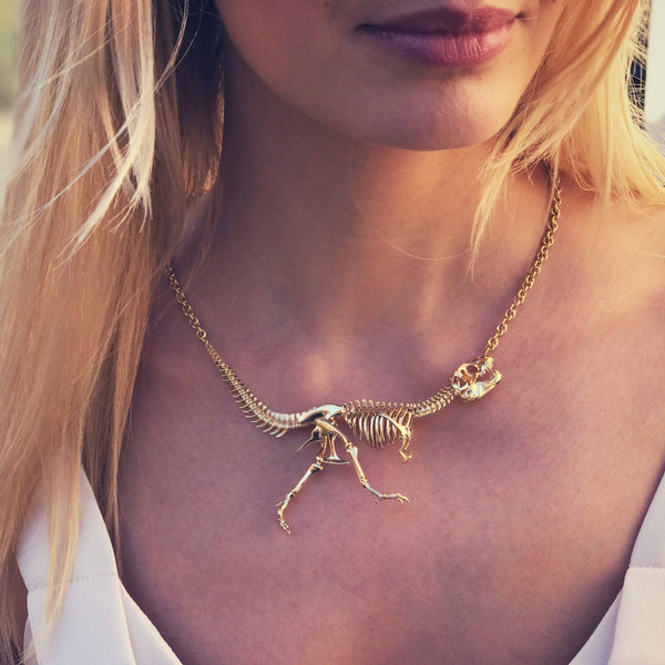 Woman wearing a yellow-gold Tyrannosaur NEX necklace—an articulated T-rex skeleton draped across her collarbone in warm summer light.